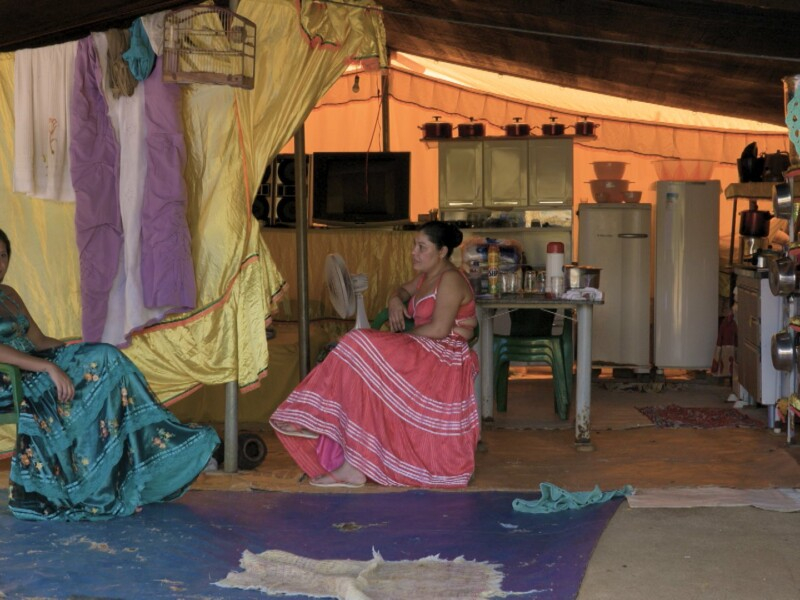 Duas mulheres estão com vestidos bem longos, um azul e outro rosa, sentadas e conversando do lado de fora do que parece ser uma cabana
