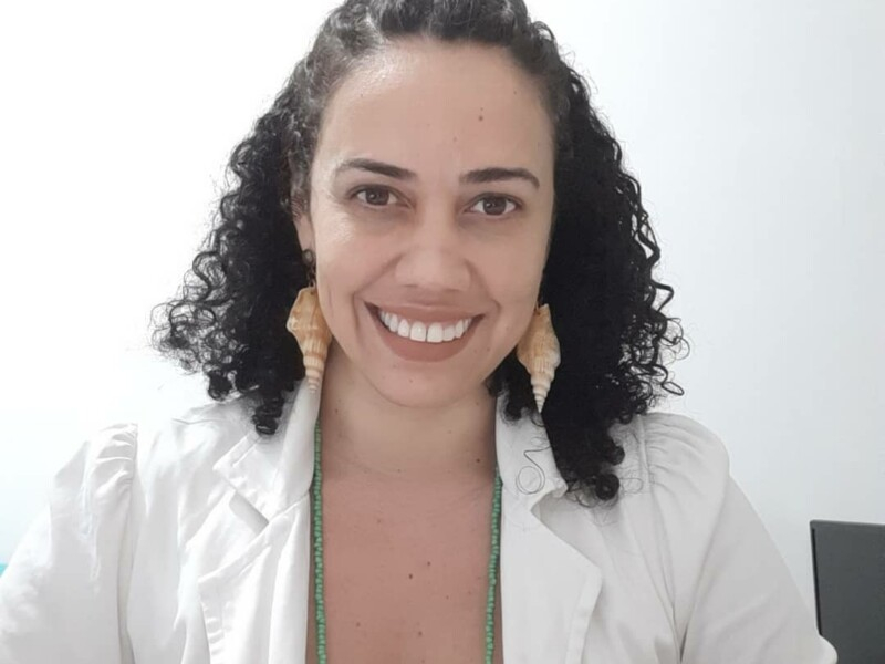 Mulher de cabelos cacheados e brincos de concha está sorrindo para quem bateu a foto
