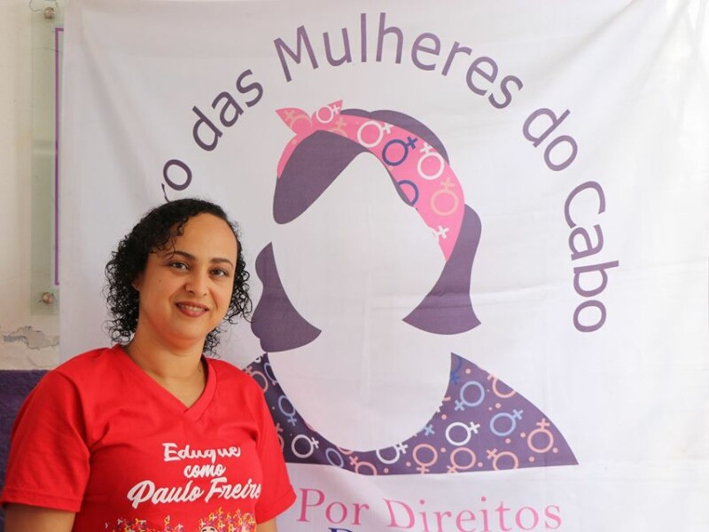 Uma mulher sorridente para quem bateu a foto está trajando uma camisa vermelha e à frente de uma bandeira branca com a logomarca do Centro das Mulheres do Cabo