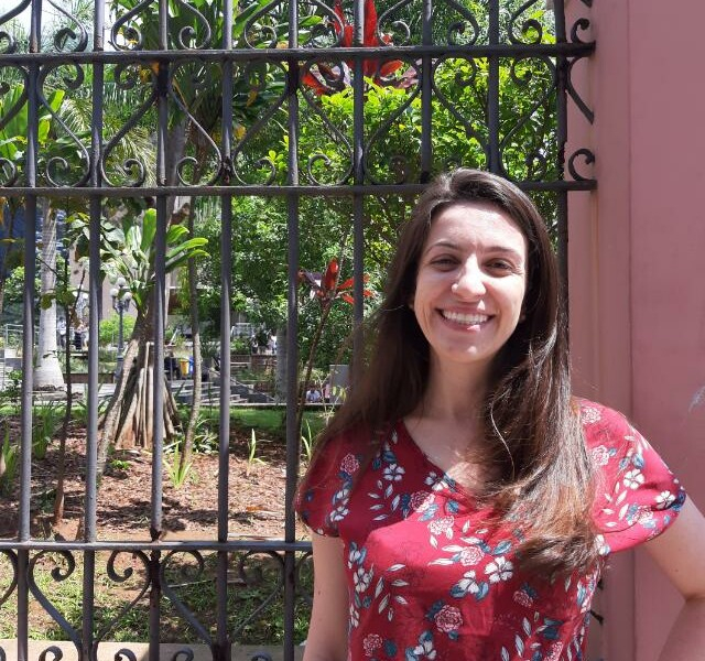Mulher branca de blusa vermelha com detalhes brancos está sorridente em frente a um portão de ferro