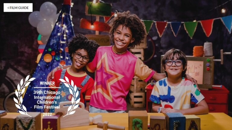 Três crianças com roupas bem coloridas estão lado a lado e posando sorridentes para a foto. Na imagem há uma arte mostrando que a série ganhou o prêmio de 1° lugar