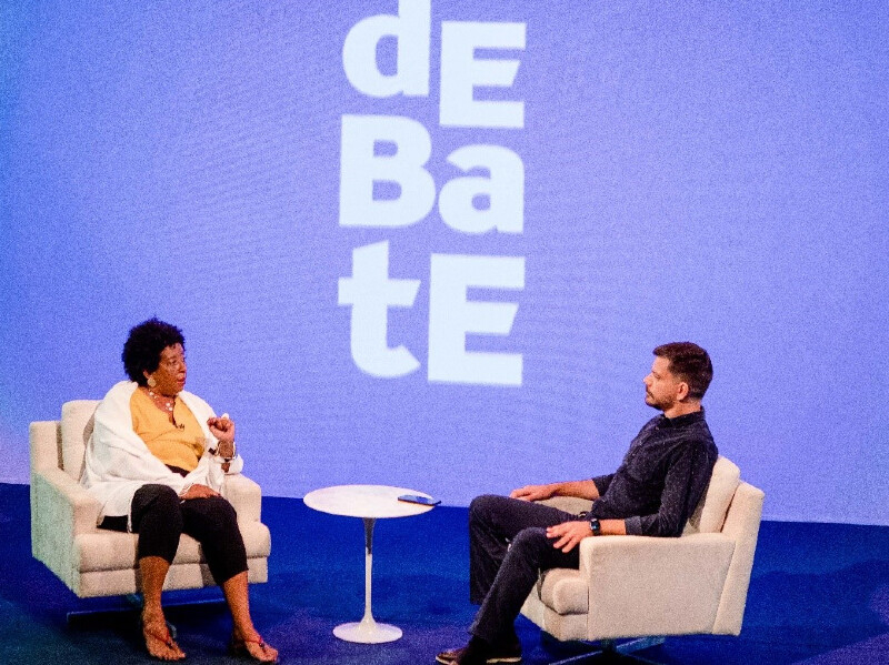 Uma mulher negra de cabelos curtos está sentada em uma poltrona frente a frente com um homem branco sentado em uma poltrona idêntica à dela. Entre eles há uma mesinha pequenina e o logotipo do programa Debate na tela azul ao fundo