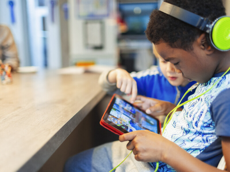 Um menino negro está de headphone e olhando atentamente para um tablet, que está em suas mãos