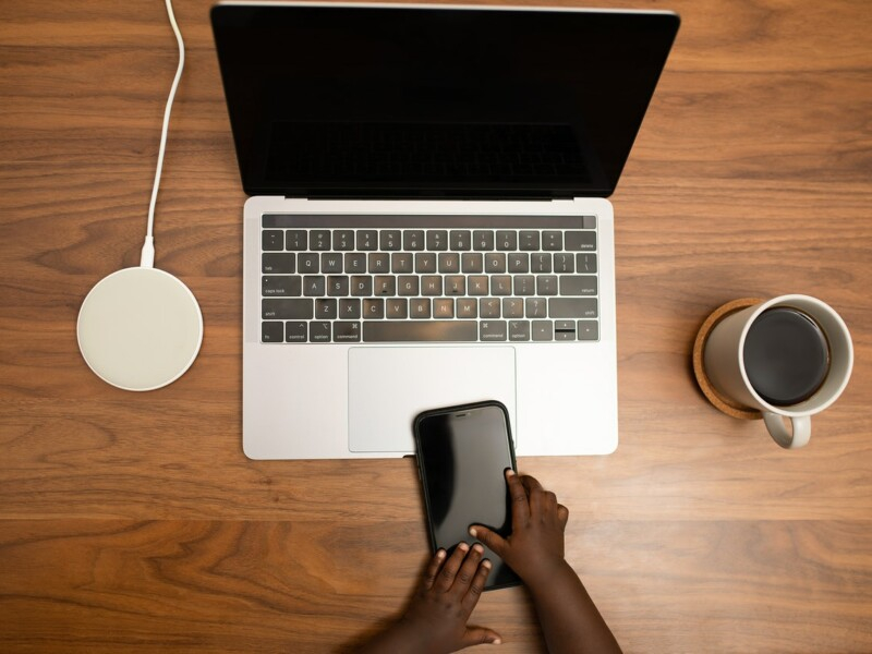Imagem aérea de um notebook aberto em cima de uma mesa de madeira. Ao lado, há uma xícara branca com um líquido preto (parece ser café) e em frente ao notebook há um celular preto sendo tocado por duas pequeninas mãos negras, que parecem ser de um bebê