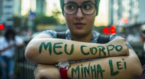 Uma mulher está ao ar livre, parece ser uma manifestação. Ela está com um braço por cima do outro posando para a foto e a frase "Meu corpo, minha lei" está escrita em seus antebraços na cor verde