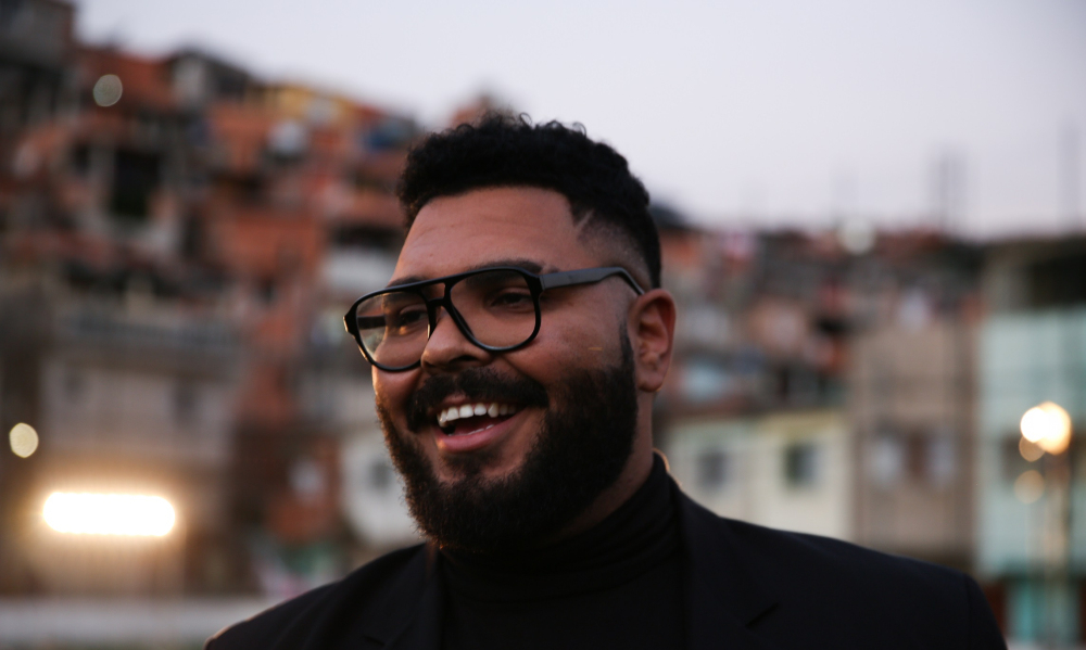Comediante Paulo Vieira está sorridente e usando óculos de grau na foto. Ao fundo podemos notar uma comunidade