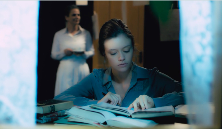 Menina branca lendo um livro em frente à uma janela. Ao fundo, há uma adulta em pé