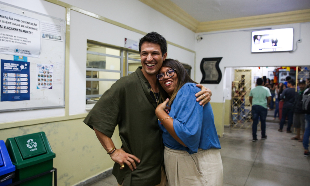 Ator Rômulo Estrela abraçando uma mulher negra de óculos
