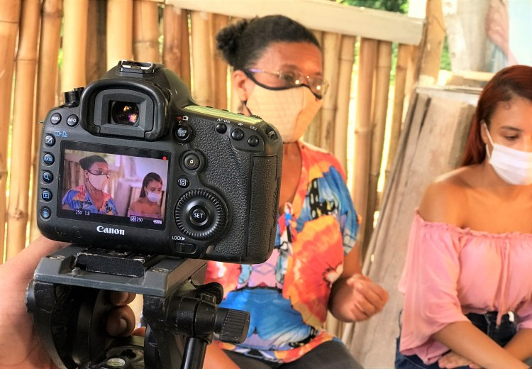 Uma máquina fotográfica está em um tripé focalizando duas mulheres de máscaras de prevenção à covid-19