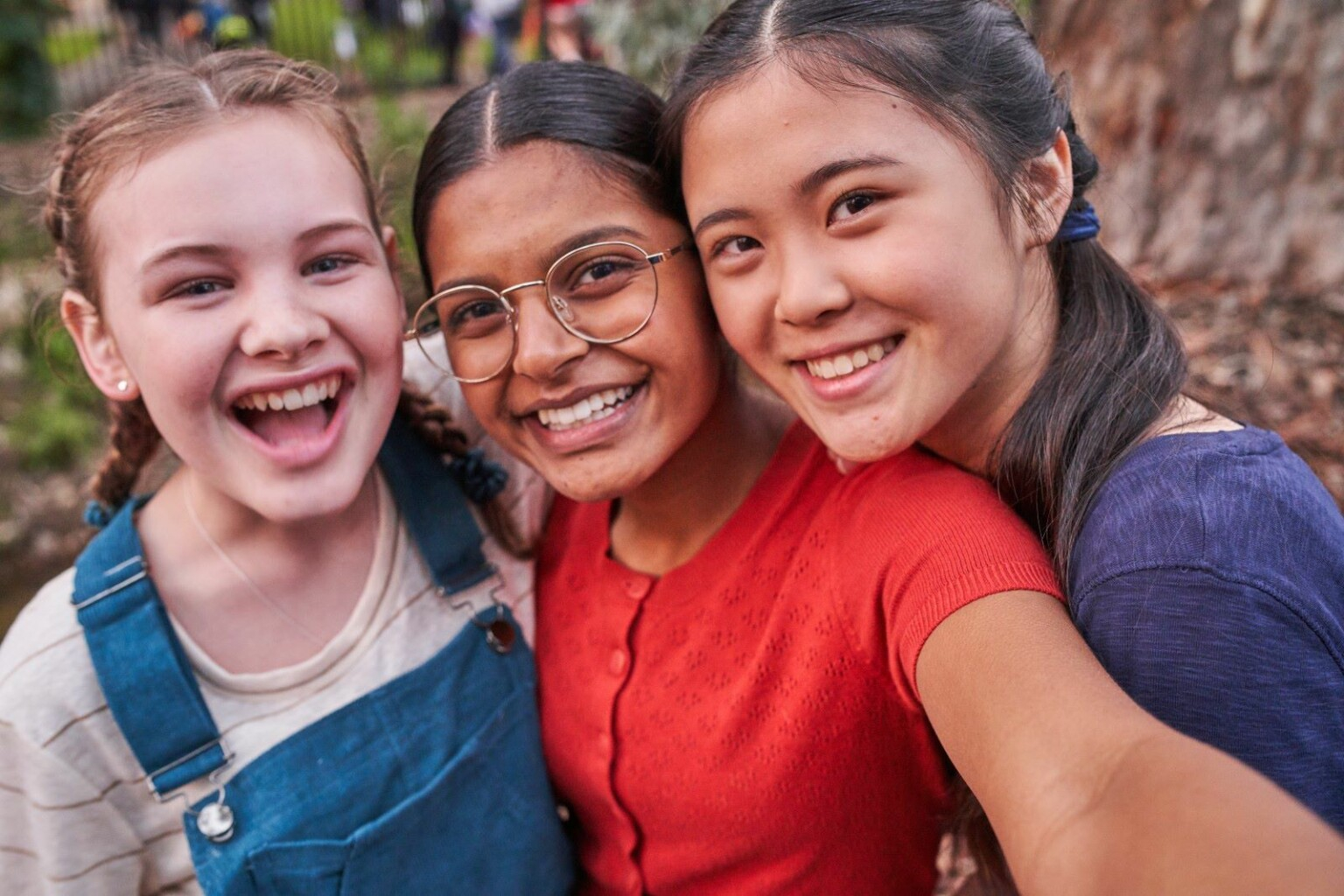 Três meninas lado a lado estão sorrindo e mostrando imensa felicidade. A foto foi uma selfie que a menina do meio fez. Uma é loira e branca, outra é negra de óculos e a outra é oriental