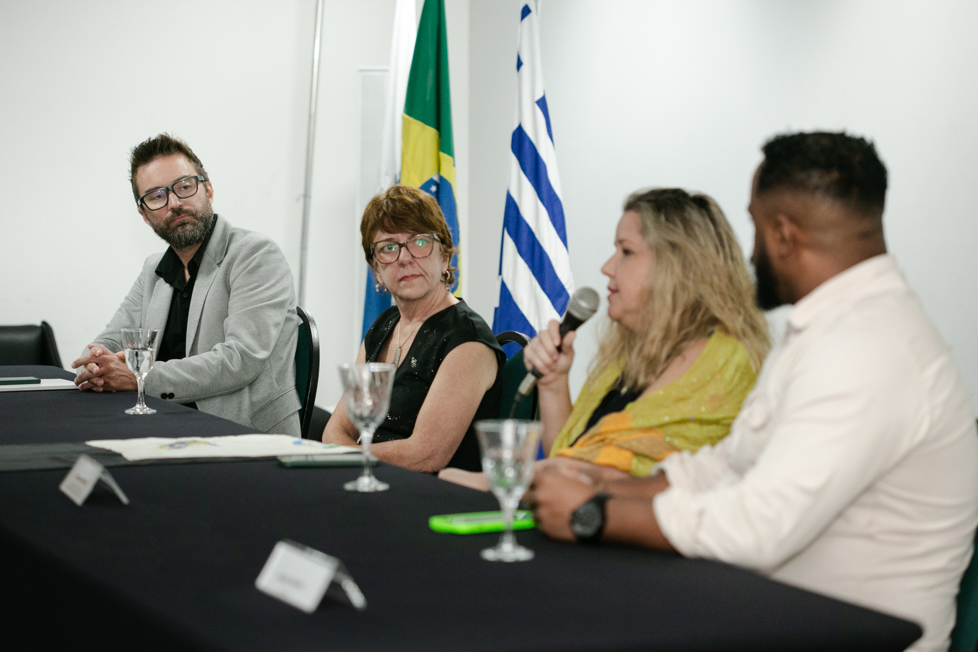 Quatro pessoas estão sentadas a uma mesa: dois homens nas pontas, duas mulheres no meio. Uma delas está falando ao microfone.