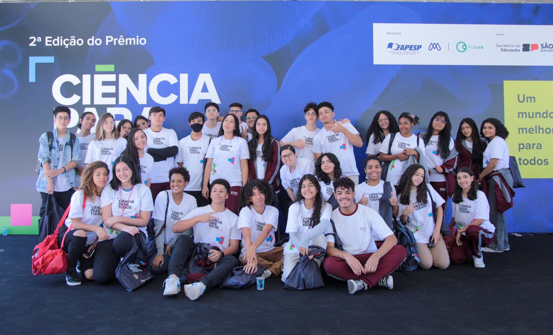 Diversas(os) jovens reunidos, de pé e sentados, sorrindo, diante de quem os fotografa. Ao fundo, um painel azul com o nome do Prêmio Ciência para Todos.