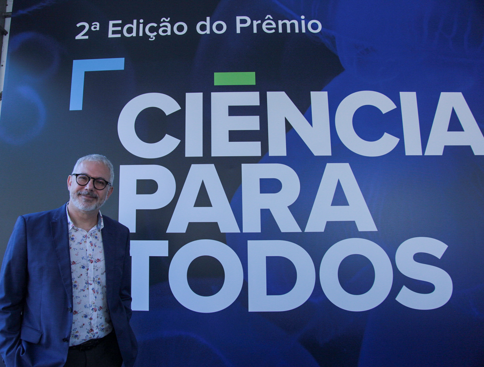 À esquerda, um homem de cabelo e barba brancos. Ao fundo, um painel azul com o nome Ciência para Todos escrito nele.