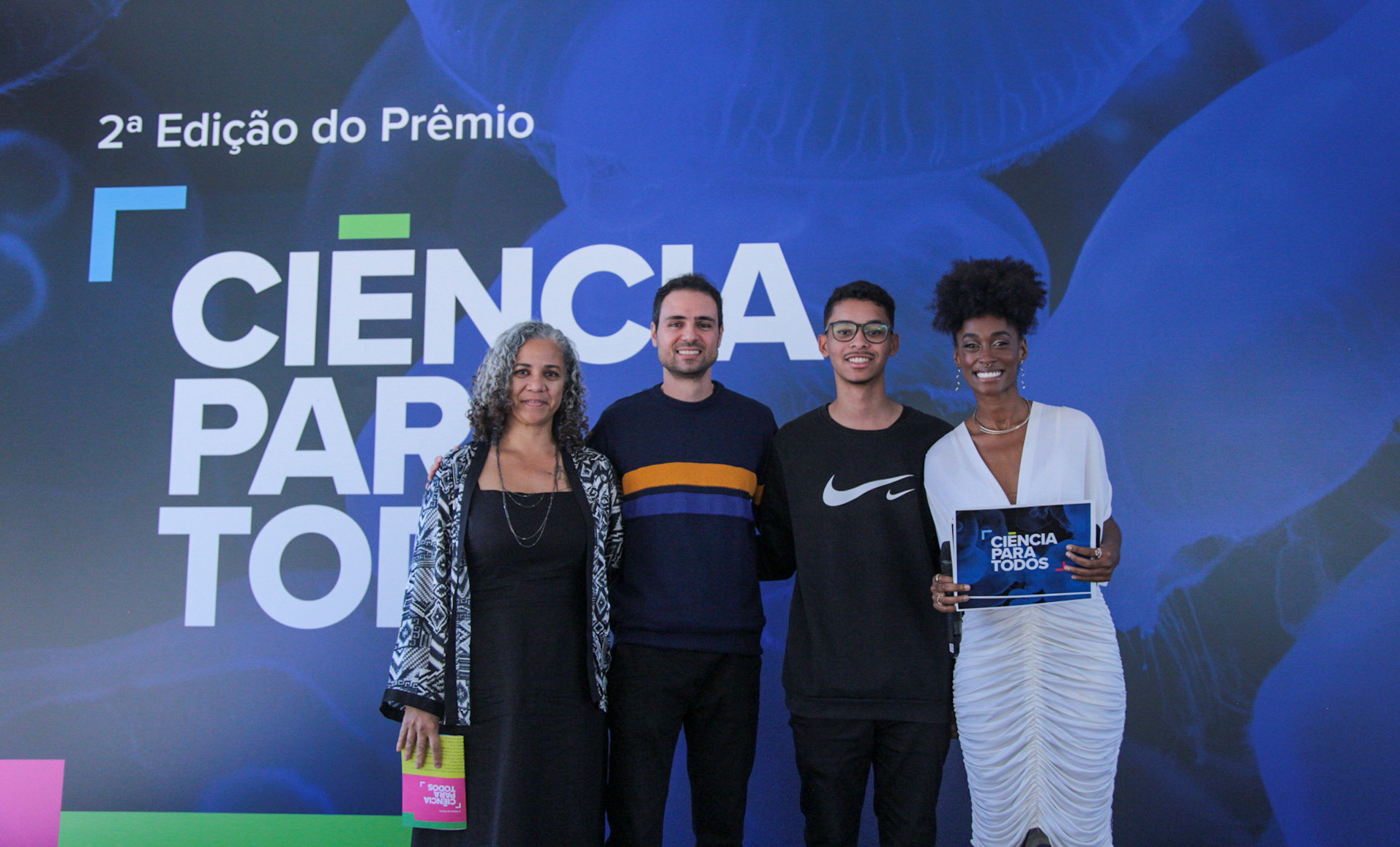 Quatro adultos - dois homens, ao centro, e duas mulheres, nas pontas -, de pé, lado a lado, sorriem para quem as fotografa.