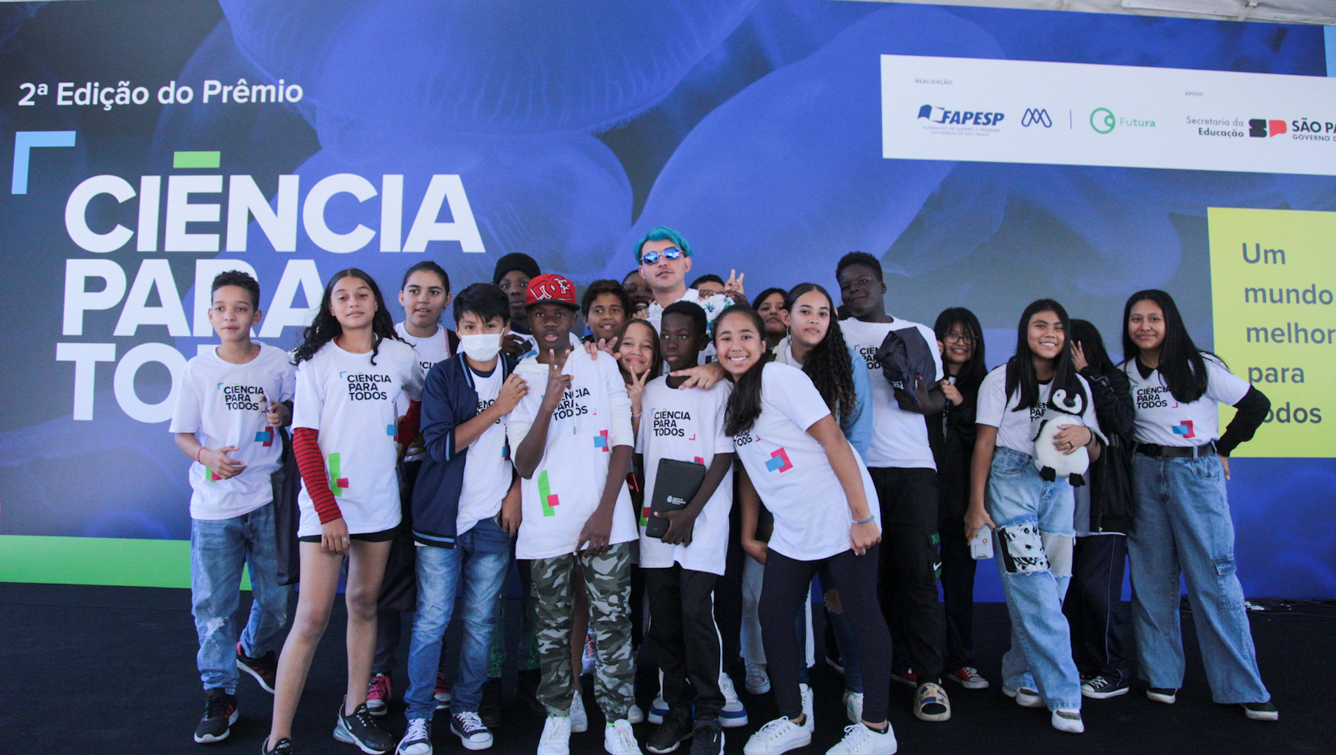 Diversos e diversas jovens reunidos de pé diante do painel com o nome do prêmio Ciência para Todos.