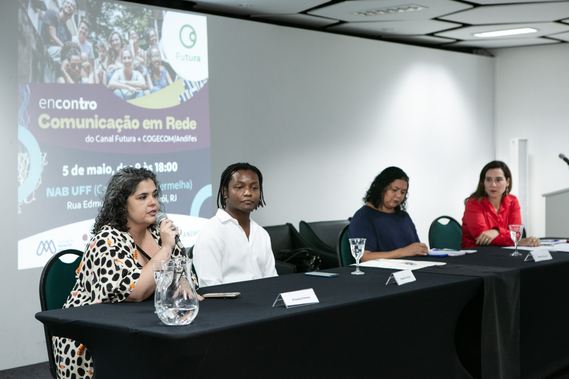 Quatro pessoas estão sentadas a uma mesa - três mulheres e um homem. Uma delas fala ao microfone.