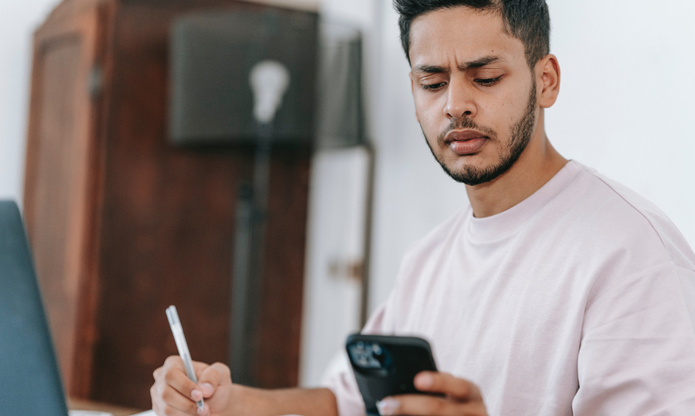 Um jovem rapaz está sentado segurando o celular com a mão esquerda e olhando para ele franzindo a testa