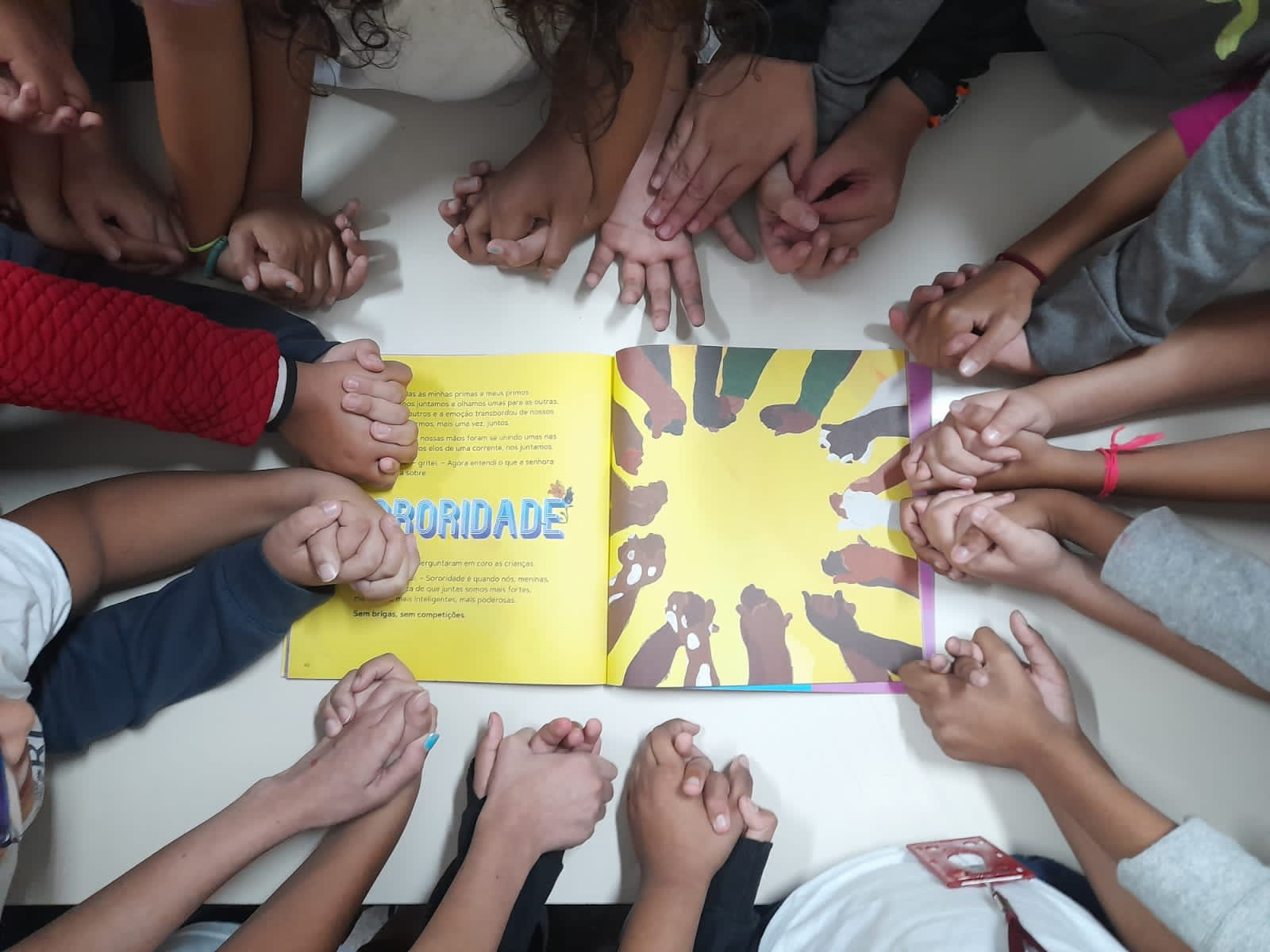 Foto de crianças dando as mãos em um livro escrito "Sororidade"
