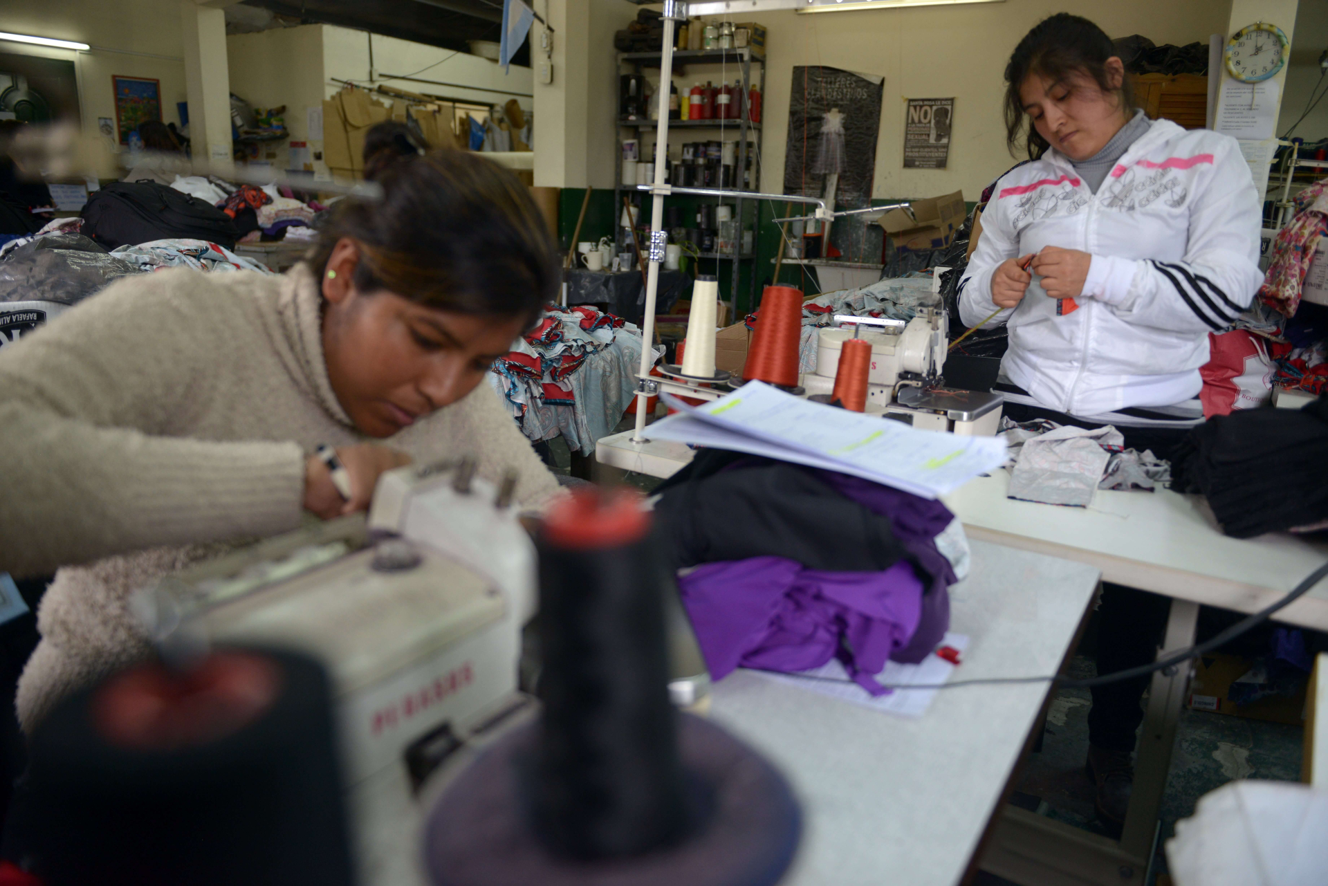 Duas mulheres mexendo em máquinas de costura e várias linhas e tecidos à sua volta