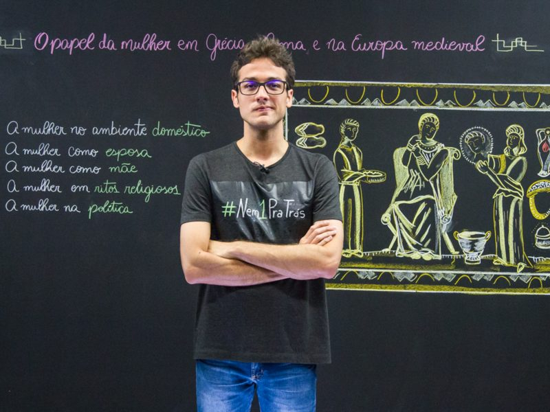 Um professor está de braços cruzados em frente ao quadro negro e posando seriamente para quem bateu a foto