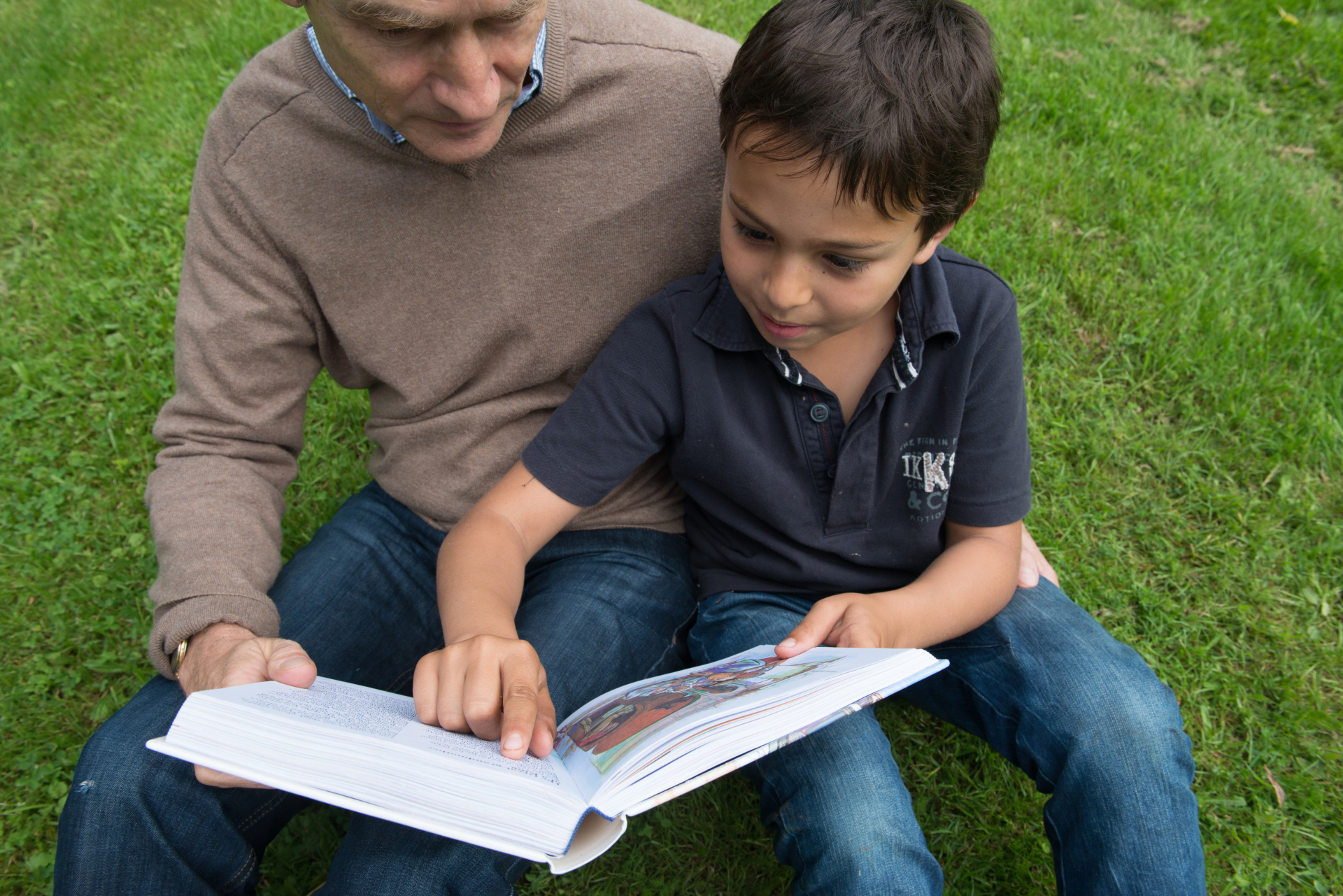 Um pai ao lado de seu filho, sentados em um gramado, olhando e lendo um livro