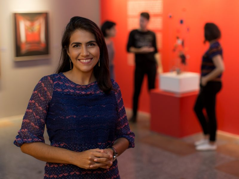Uma mulher morena está sorrindo para quem tirou a foto e está em uma galeria de artes