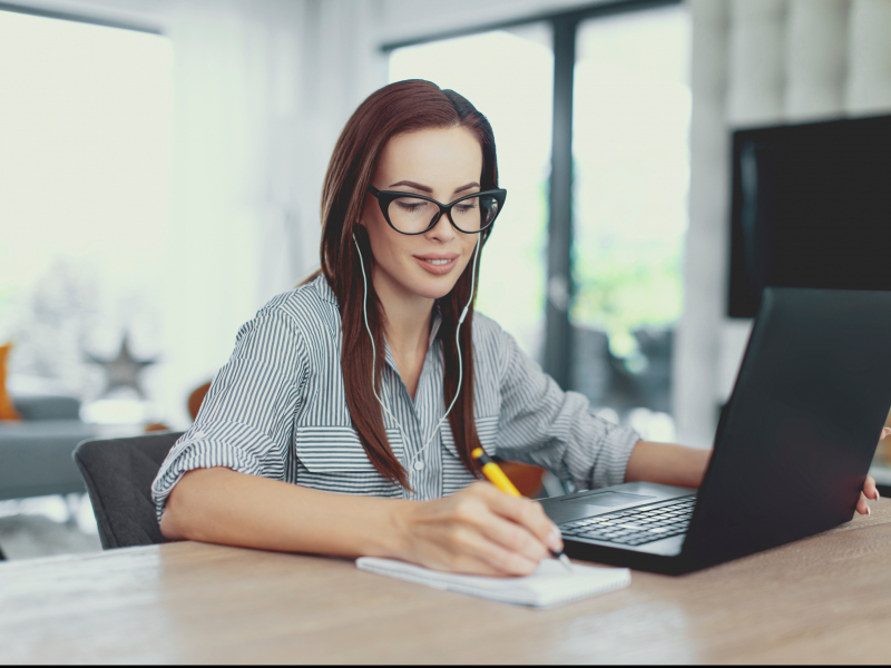 Uma jovem mulher branca e de óculos de grau e armação preta está sentada diante de um notebook aberto, enquanto escreve algo com a mão direita em um bloquinho e está de fones de ouvido