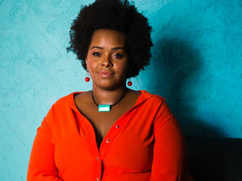 A professora Ellen Ferreira está posando para a foto. Ela ´´e uma mulher negra, de cabelo black power e está com brincos vermelhos, uma blusa laranja de botões e um belo cordão