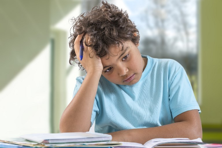 Um menino com a mão na cabeça e olhando para um ponto avulso parece preocupado com o estudos. Ele está sentado diante de livros e um caderno aberto