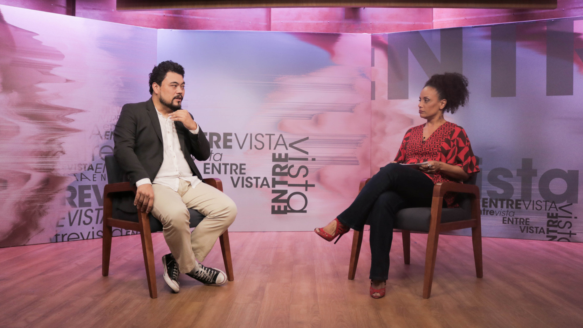 Homem oriental de paletó está sentado em uma cadeira e sendo entrevistado por uma mulher negra à sua frente, que é a apresentadora do programa ENTREVIST Flavia Oliveira