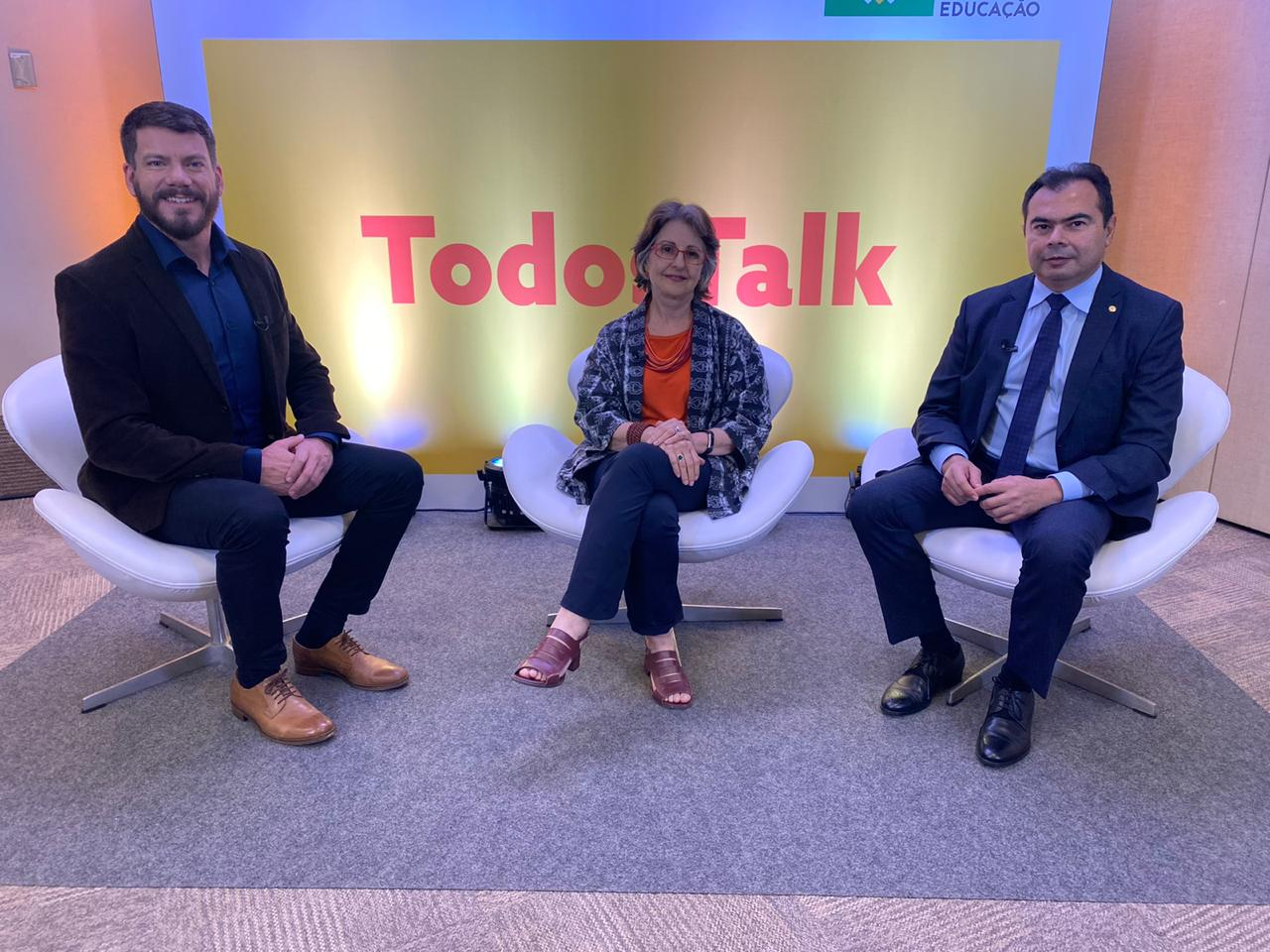 Cenário do programa Debate com o apresentador e dois convidados sentados em cadeiras brancas