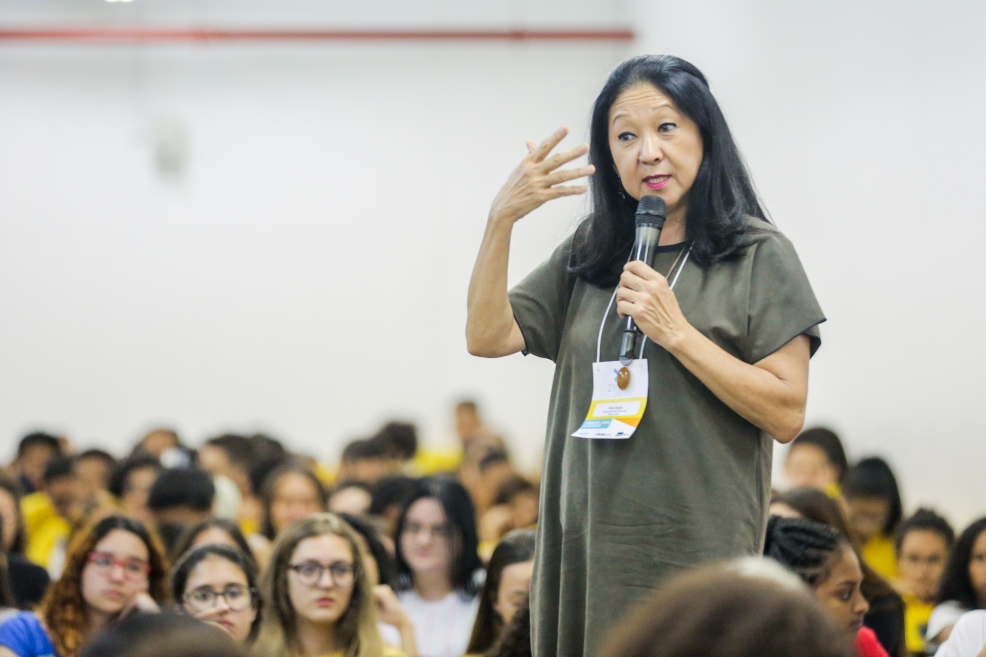 Mulher oriental está em pé falando em um microfone, meio a várias pessoas sentadas. Trata-se de uma plateia.