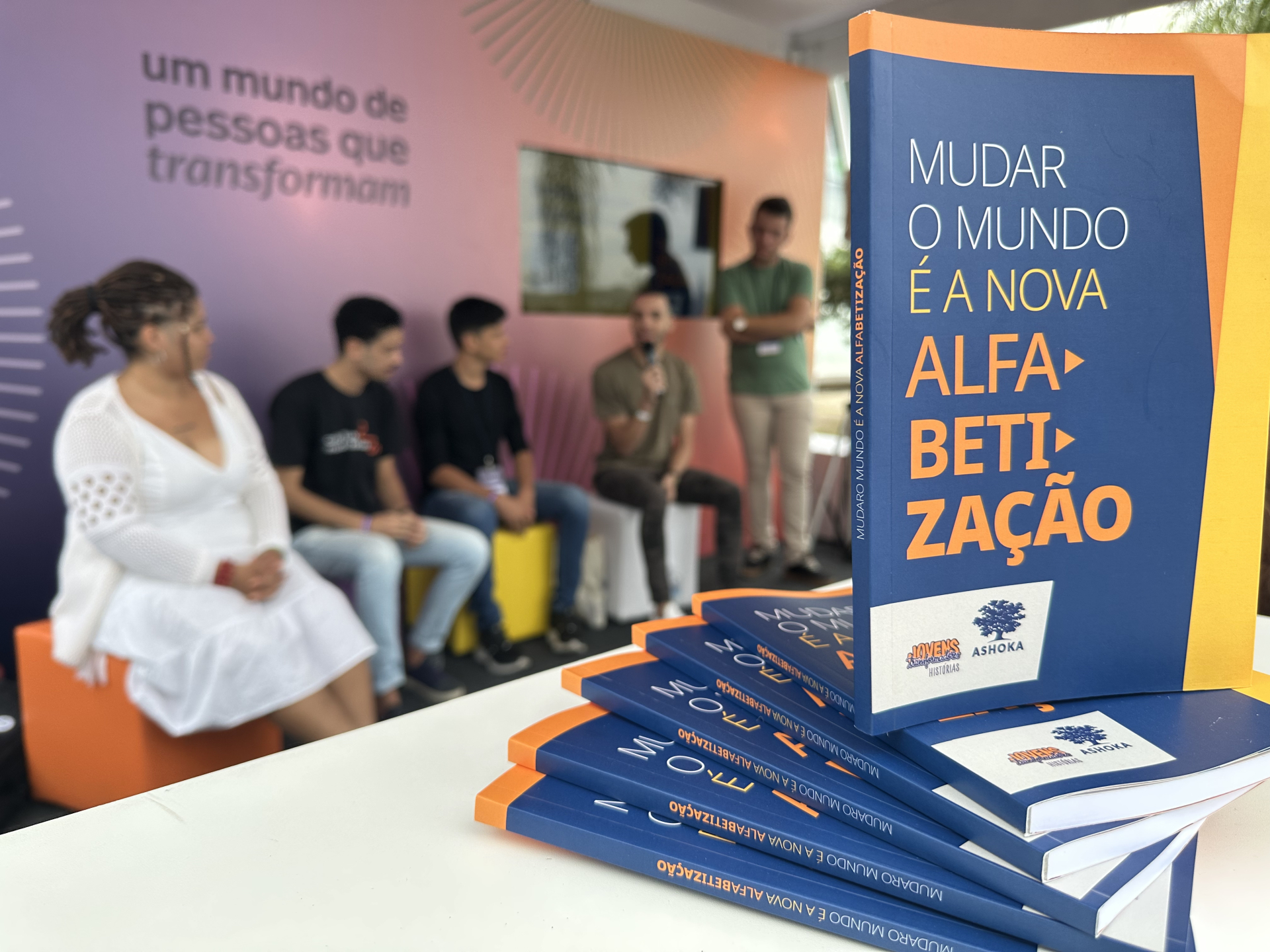 Foto mostra, em primeiro plano, a capa do livro "Mudar o mundo é a nova alfabetização". Ao fundo, jovens sentados em poltronas coloridas.