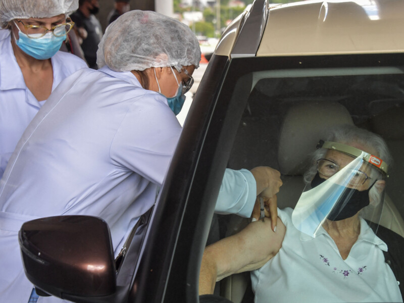 Uma pessoa da área da saúde está aplicando uma vacina no braço direito de uma senhora, que está sentada dentro de um carro e usando máscara de prevenção à covid
