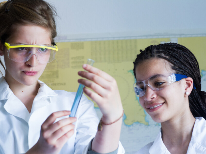 Duas jovens mulheres de jaleco e óculos de proteção estão em um possível laboratório manuseando seringas