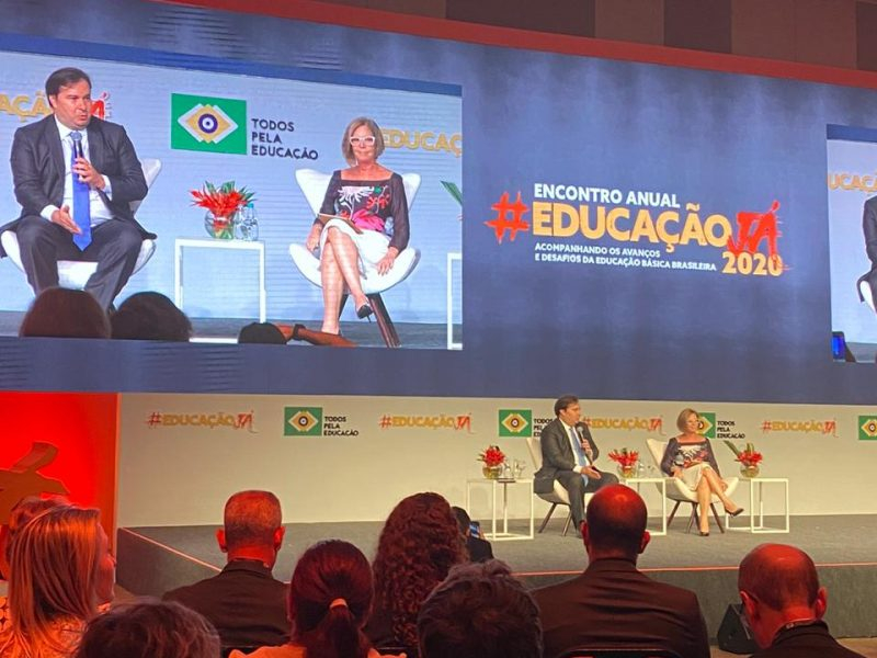 Um homem de terno e uma mulher bem vestida estão sentados em cadeiras em cima de um palco. Eles estão com microfones e falando algo para uma grande plateia