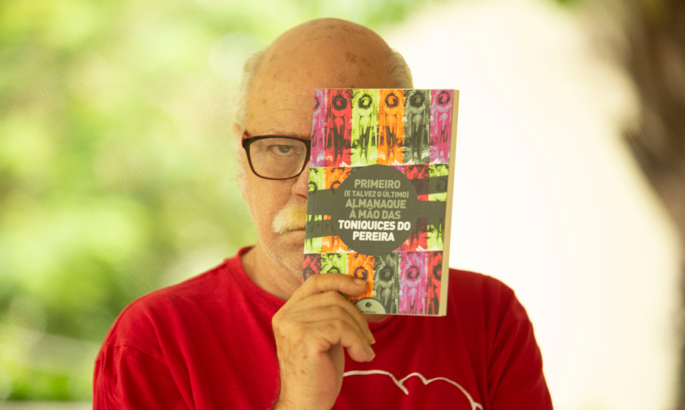 Ator idoso Tonico Pereira está segurando um livro em frente à metade esquerda de sua face e posando seriamente para a foto