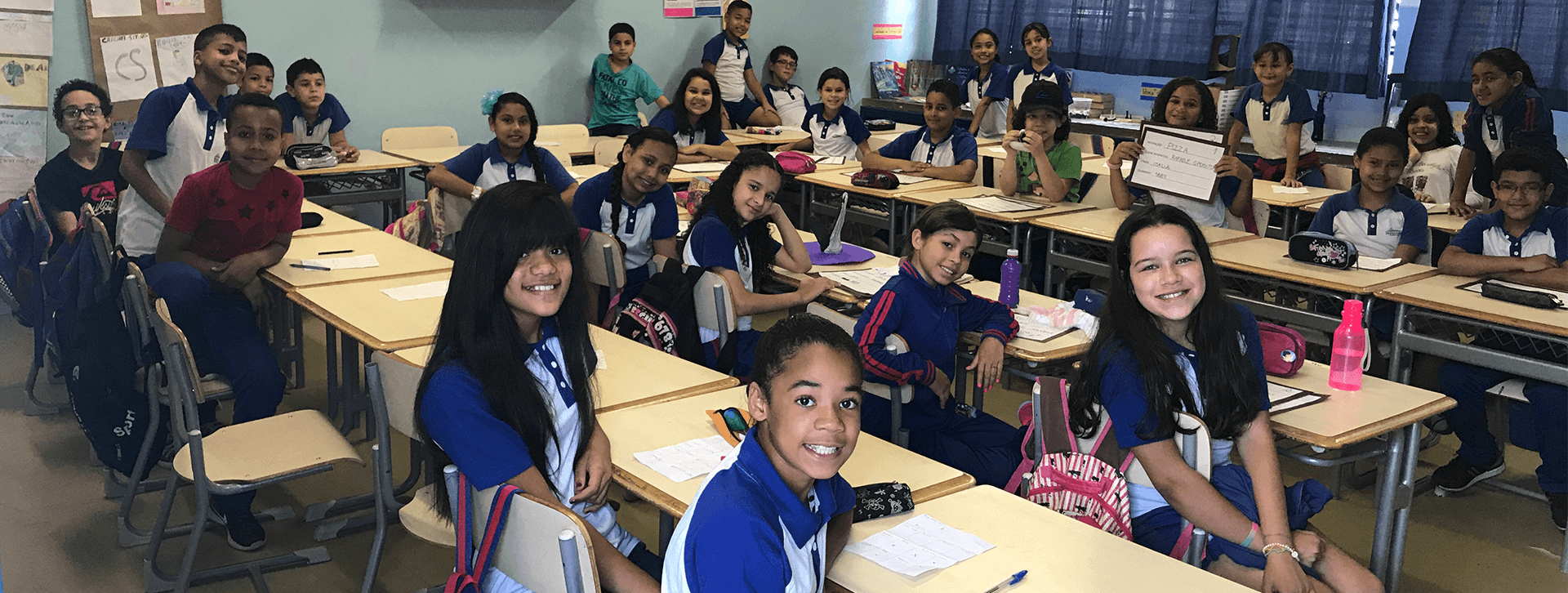 Uma sala de aula com vários alunos olhando para quem bateu a foto e sorrindo