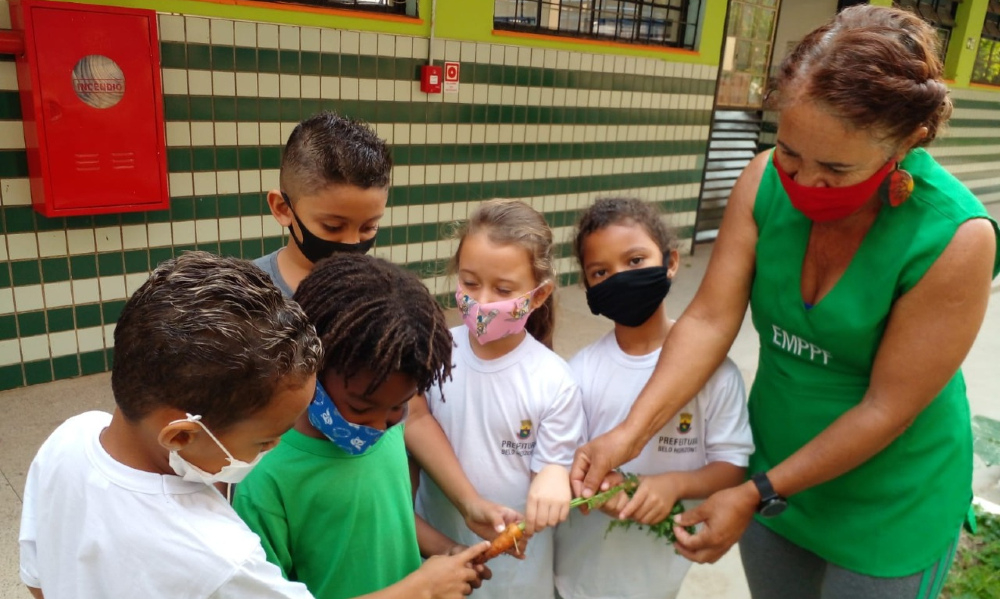 Uma mulher adulta está mostrando uma cenoura para cinco crianças. Todas as pessoas estão usando máscara de proteção à covid