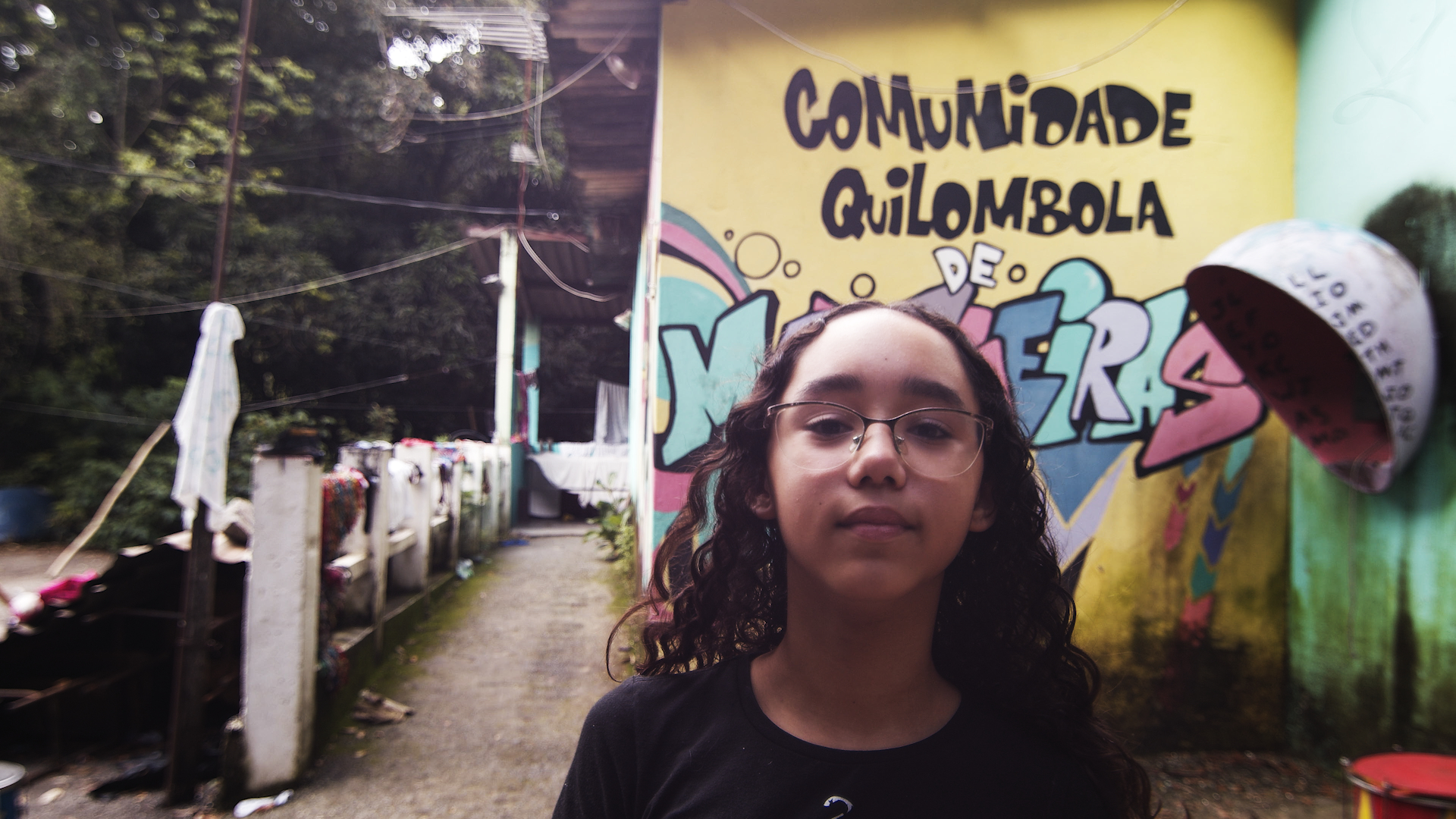Uma jovem, usando óculos, está olhando para quem a fotografa. Atrás dela, uma parede com o texto escrito em preto: "Comunidade quilombola".