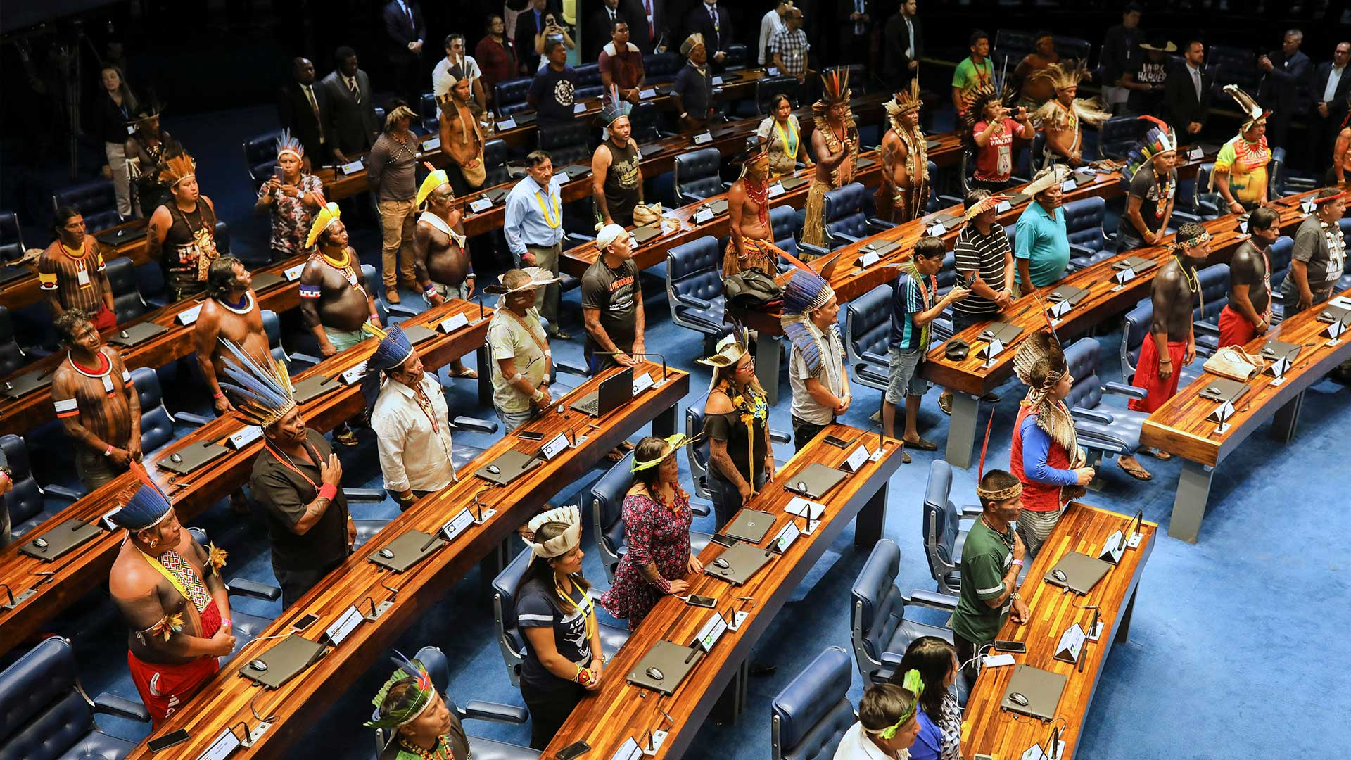 Dezenas de indígenas estão em um local que parece ser o congresso, todos em pé, atrás de bancadas