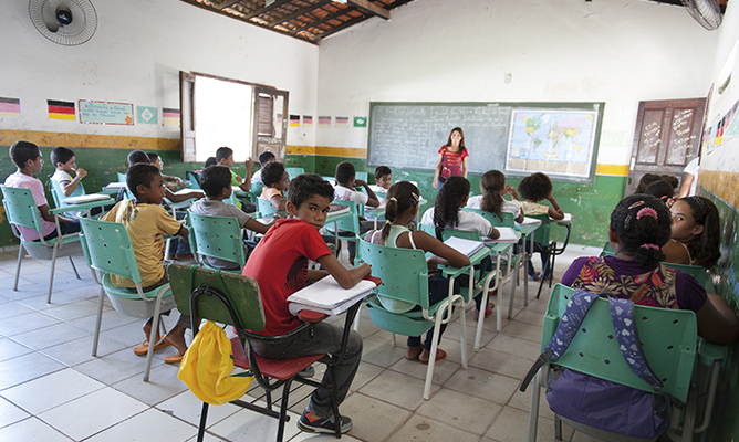 Uma sala de aula com vários alunos sentados prestando atenção na professora em pé falando