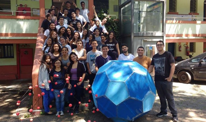 Vários alunos estão em pé em uma escadaria e à frente deles há um professor e uma grande bola azul