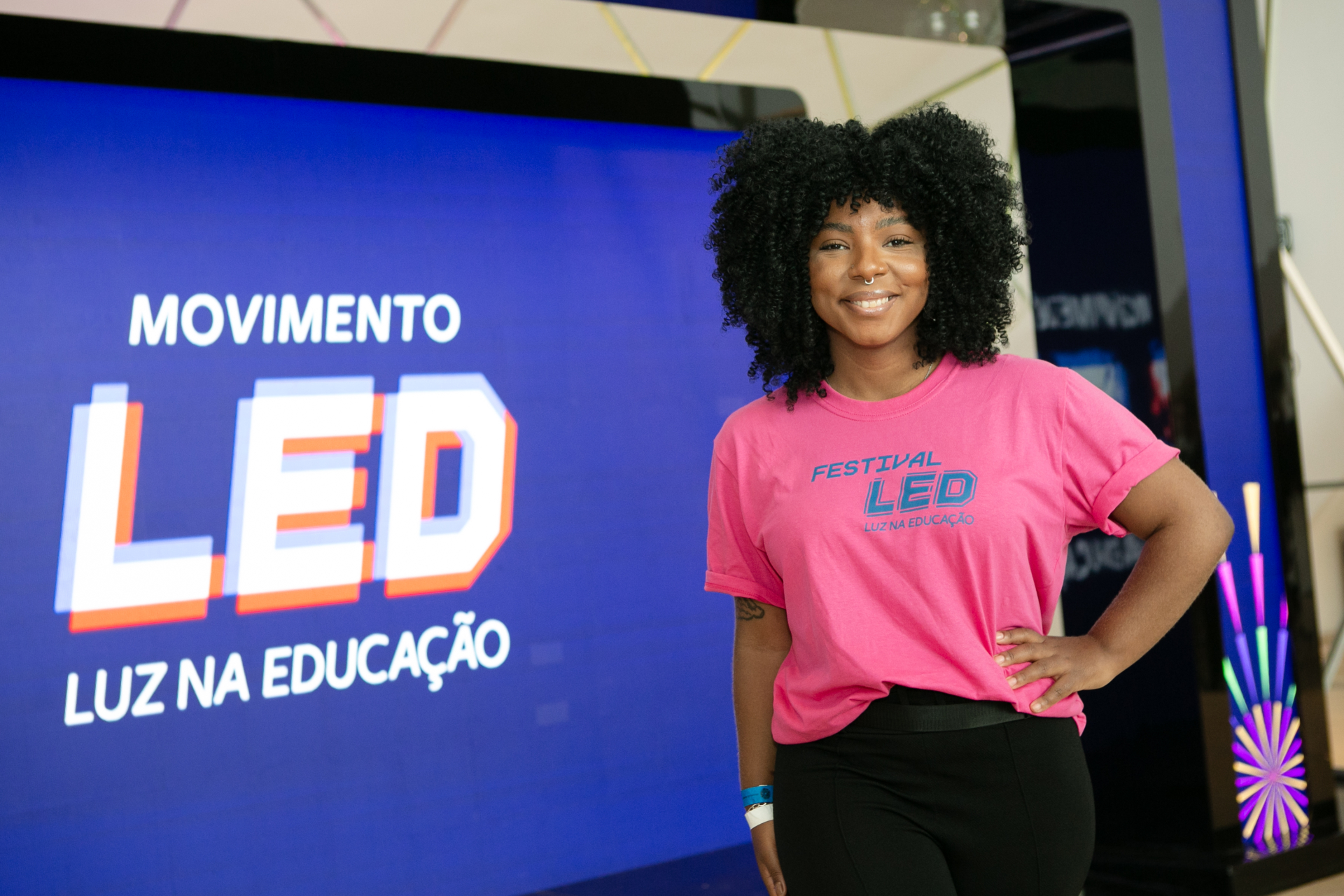 Uma mulher negra sorri ao lado de um painel com o texto "Movimento LED - Luz na Educação".