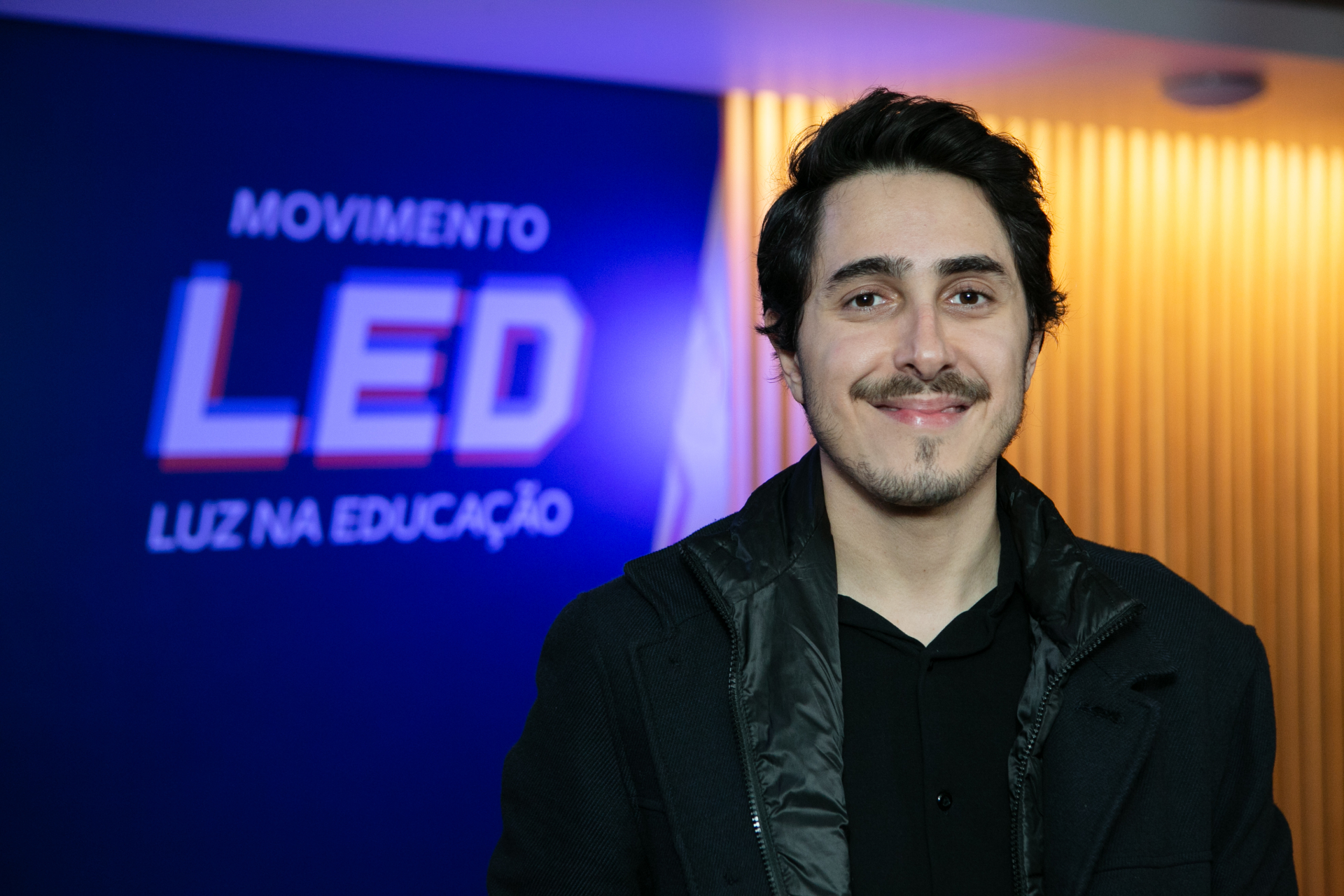 Um homem, com bigode e cavanhaque, sorri. Ao fundo, um painel em tons de azul com o texto "Movimento LED - Luz na Educação".