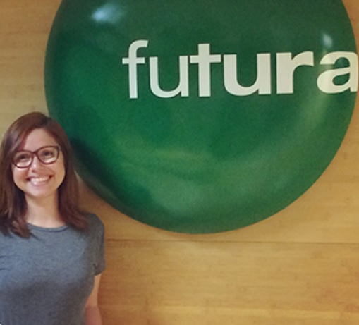 Jovem mulher branca, de óculos de grau e cabelos lisos avermelhados está sorridente para quem bateu a foto e posando à frente da logomarca do Canal Futura