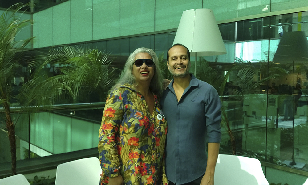 Uma mulher de cabelos grisalhos e soltos está ao lado de um homem calvo de camisa social. Ambos estão posando sorridentes para quem bateu a foto