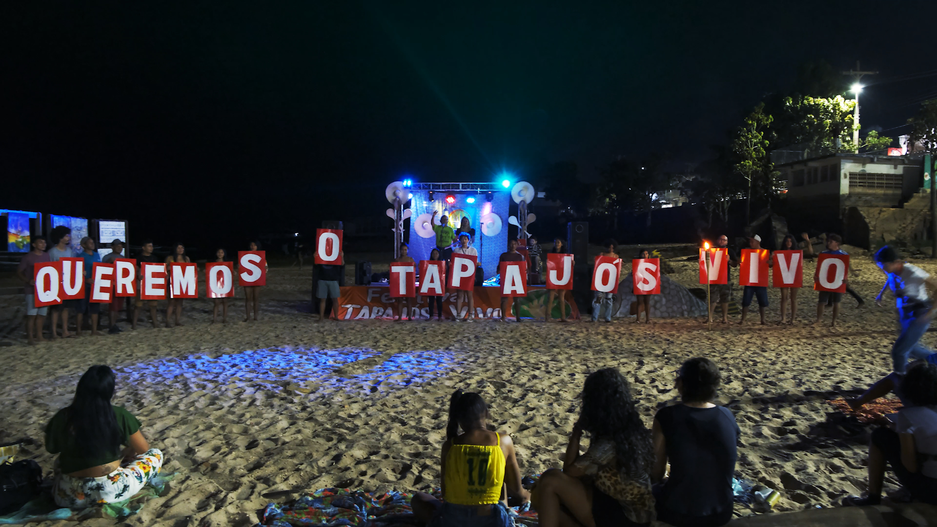 Jovens estão sentados na areia, diante de cartazes que têm o seguinte texto: "Queremos o Tapajós vivo".