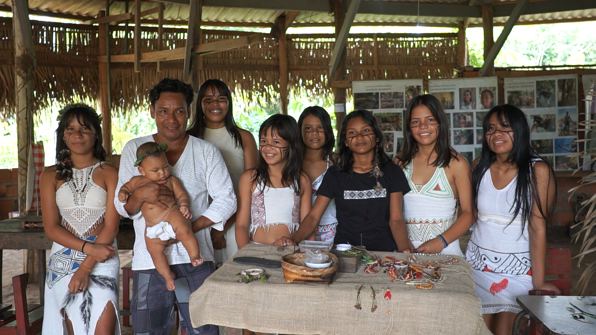 Diversos jovens indígenas estão reunidos e reunidas diante de uma mesa, sob um teto de palha.