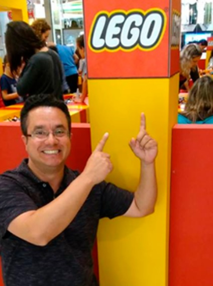Um professor de óculos está agachado e apontando para cima, onde tem a logomarca da Lego